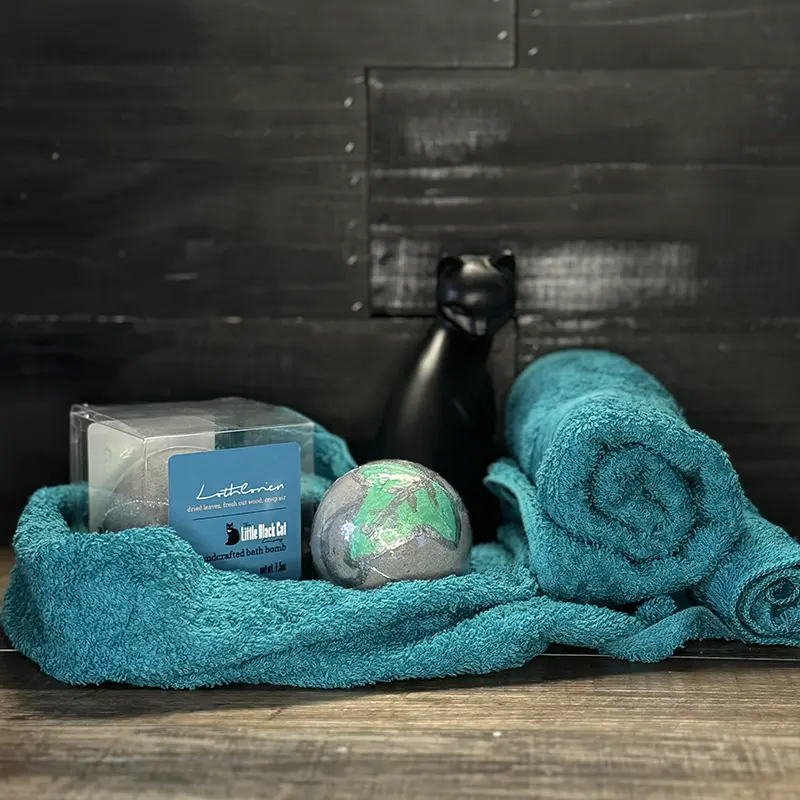 artistic arrangement of one boxed and one loose bath bomb with a towel and an art deco cat statue